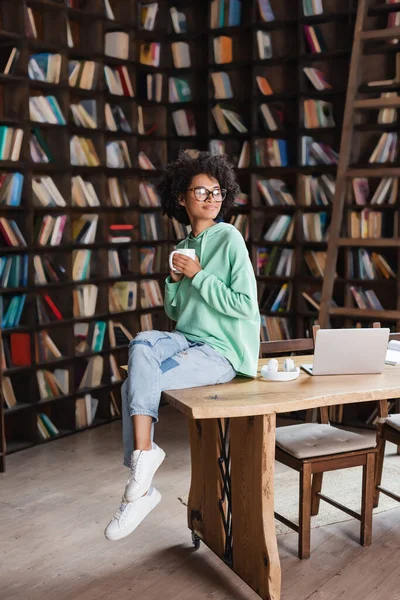 Ricci donna afro-americana in occhiali con in mano una tazza di caffè vicino al computer portatile sulla scrivania — Foto stock