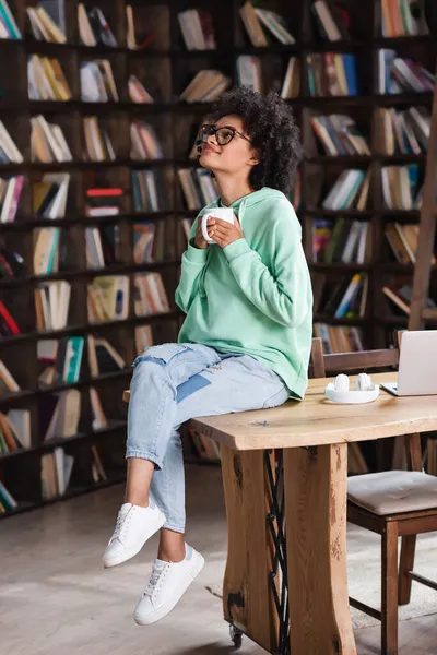 Piacere afro americano donna in occhiali in possesso di una tazza di caffè vicino al computer portatile sulla scrivania — Foto stock