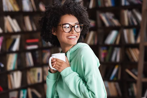 Счастливая молодая африканская американка в очках держит чашку кофе — стоковое фото