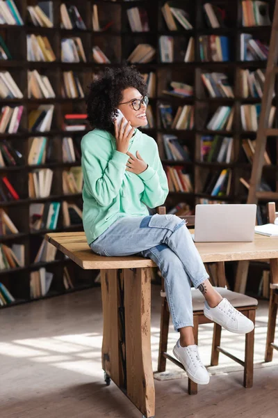 Gioiosa donna afro-americana con gli occhiali che parla sul cellulare vicino al portatile sulla scrivania — Foto stock