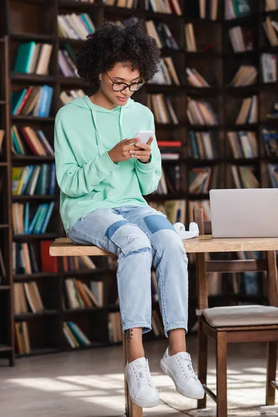 Giovane donna afroamericana in occhiali da vista utilizzando il cellulare mentre seduto sulla scrivania vicino al computer portatile — Foto stock