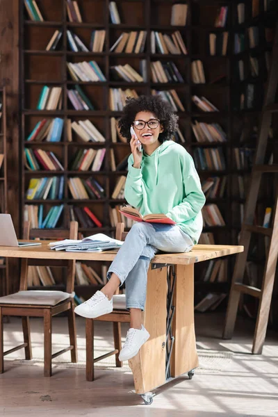 Fröhliche afrikanisch-amerikanische Studentin in Brille, die auf dem Handy neben Laptop und Notebooks auf dem Schreibtisch spricht — Stockfoto