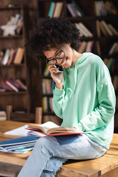 Alegre africano americano estudiante en gafas hablando en el teléfono celular y lectura libro y sentado en escritorio - foto de stock