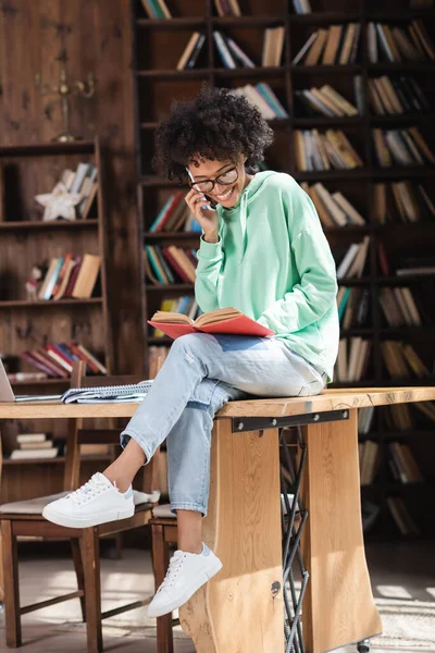 Felice studente afroamericano con gli occhiali che parla sul cellulare e legge un libro mentre è seduto sulla scrivania — Foto stock