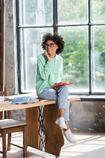 Happy african american student in eyeglasses talking on cellphone and holding book while sitting on desk — Stock Photo
