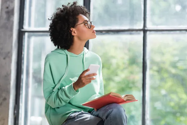 Curly african american student in eyeglasses holding cellphone and book while looking at window — Stock Photo