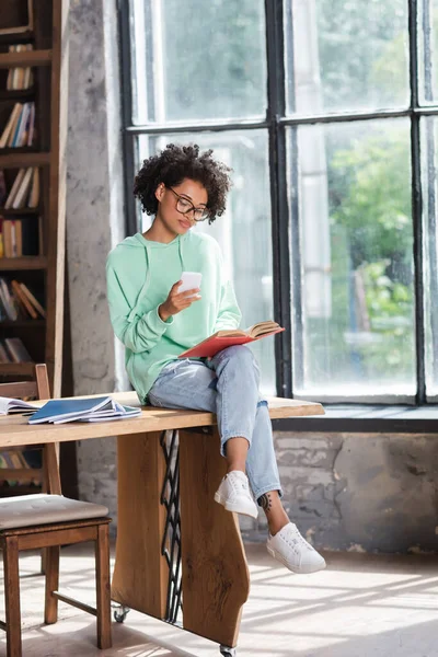 Lockige afrikanisch-amerikanische Studentin mit Brille, Handy in der Hand und Buch lesen, während sie am Tisch sitzt — Stockfoto