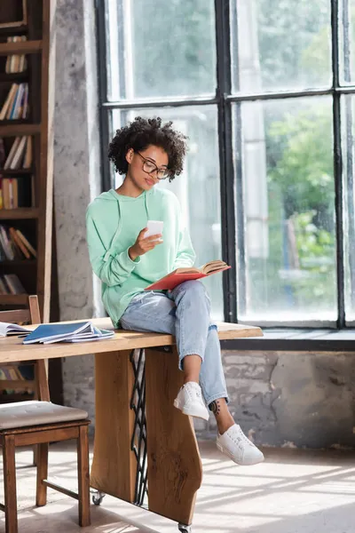 Lockige afrikanisch-amerikanische Studentin mit Brille, Handy in der Hand und Buch lesen, während sie am Tisch sitzt — Stockfoto