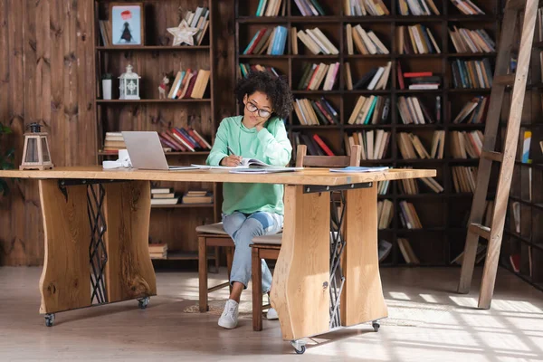 Curly african american student in eyeglasses writing in notebook near laptop — Stock Photo