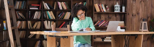 Étudiant afro-américain bouclé en lunettes écriture dans un carnet tout en étudiant en ligne près d'un ordinateur portable, bannière — Photo de stock