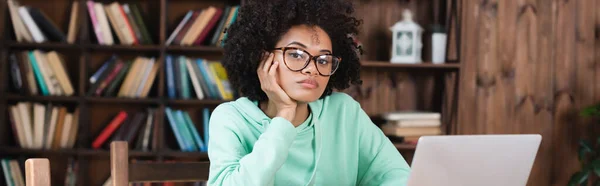 Estudiante afroamericano aburrido en gafas mirando a la cámara mientras estudia en línea cerca de la computadora portátil, pancarta - foto de stock