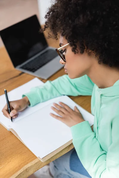 Estudiante afroamericano rizado en anteojos escribiendo en cuaderno mientras estudia en línea cerca de computadora portátil borrosa con pantalla en blanco - foto de stock