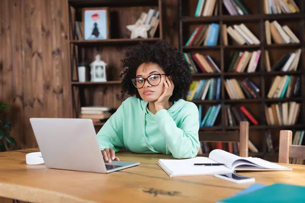 Gelangweilte afrikanisch-amerikanische Studentin mit Brille blickt während ihres Online-Studiums in der Nähe von Gadgets und Notebooks in die Kamera — Stockfoto