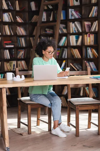 Studente afroamericano in occhiali utilizzando smartphone vicino laptop e cuffie sulla scrivania — Foto stock