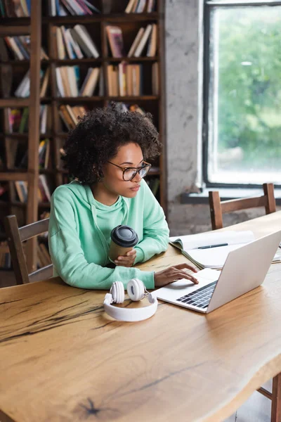 Estudiante afroamericano en gafas usando portátil mientras sostiene la taza de papel cerca de los auriculares en el escritorio - foto de stock