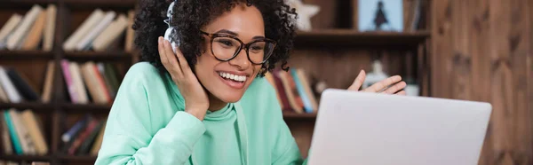 Estudiante afroamericano feliz en gafas y auriculares usando el ordenador portátil, bandera - foto de stock