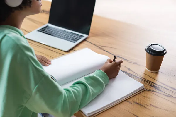 Vista recortada de estudante americano africano escrevendo em notebook perto de laptop com tela em branco — Fotografia de Stock