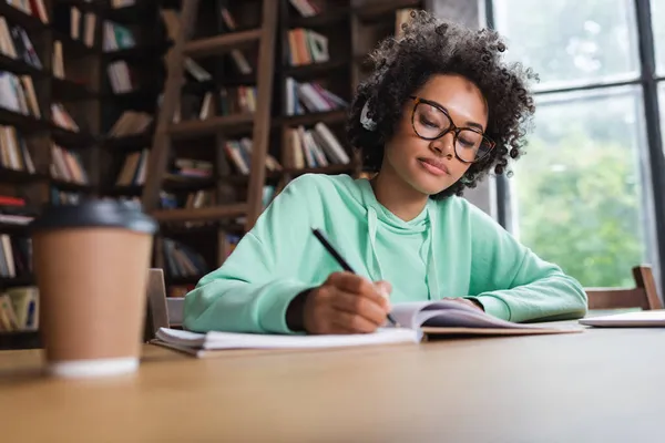 African american student in eyeglasses and headphones writing near laptop and paper cup — Stock Photo