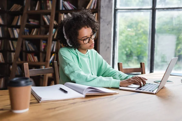 African american student in eyeglasses and headphones typing on laptop and paper cup and notebook — Stock Photo