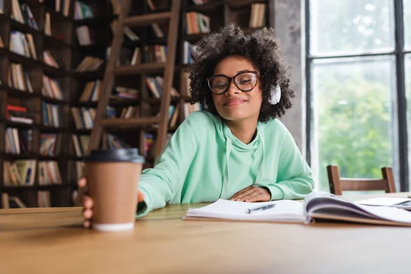 Happy african american student in eyeglasses and headphones reaching blurred paper cup — Stock Photo