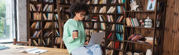 Riccio studente afroamericano in occhiali da vista in possesso di tazza di carta durante l'utilizzo di laptop, banner — Foto stock