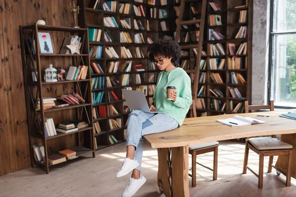 Curly african american student in eyeglasses holding paper cup while using laptop — Stock Photo