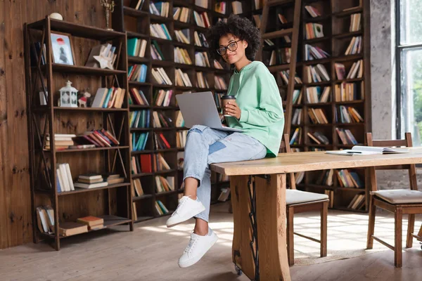 Étudiant afro-américain souriant dans des lunettes tenant tasse en papier tout en étant assis avec ordinateur portable sur le bureau — Photo de stock