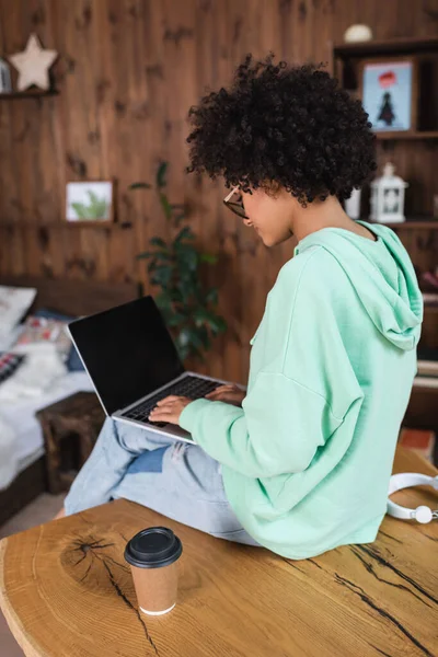 Afro-americano estudante em óculos digitando no laptop com tela em branco e sentado na mesa — Fotografia de Stock