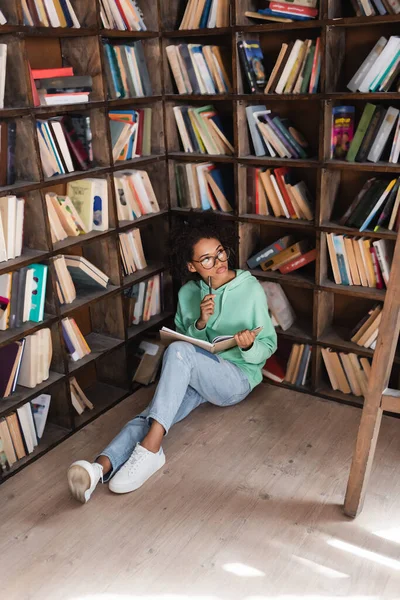 Pensive african american student in eyeglasses sitting on floor surrounded by books and holding notebook in library — Stock Photo