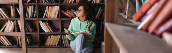 Étudiant afro-américain coûteux en lunettes assis et tenant un cahier dans la bibliothèque, bannière — Photo de stock