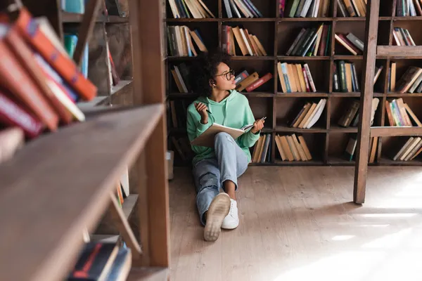 Pensive african american student in eyeglasses sitting and holding notebook in library with blurred bookshelf — Stock Photo