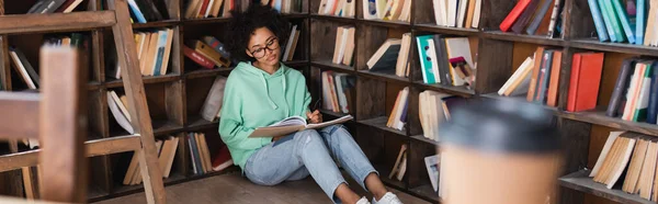 Jeune étudiant afro-américain en lunettes écrivant sur un carnet près d'une tasse en papier floue au premier plan, bannière — Photo de stock