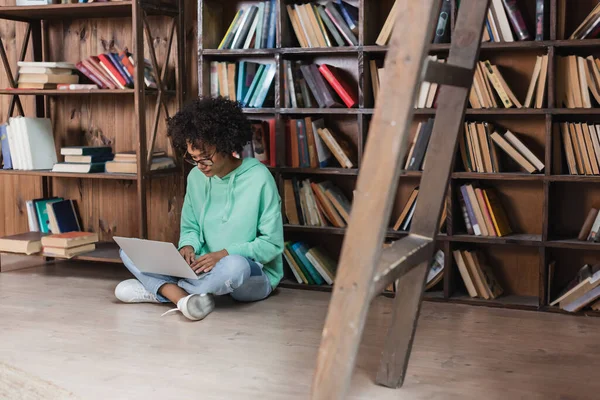 Curly african american student in eyeglasses using laptop while sitting on floor in library — Stock Photo