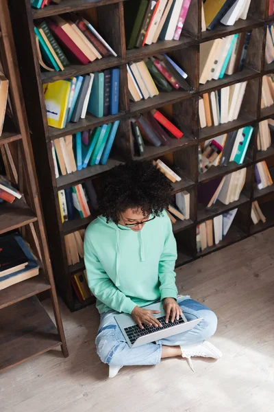 High angle view of curly african american student in eyeglasses using laptop while sitting on floor in library — Stock Photo