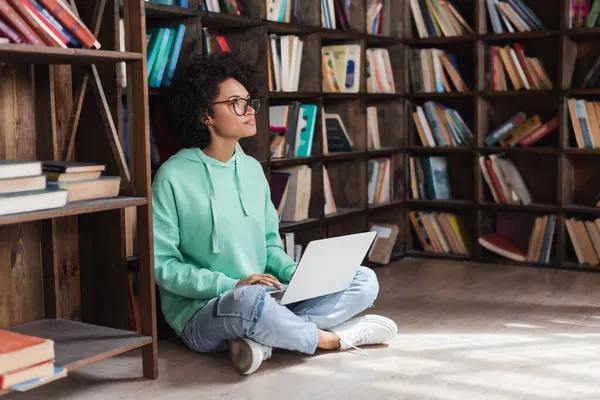 Curly african american woman in eyeglasses using laptop while sitting on floor in library — Stock Photo