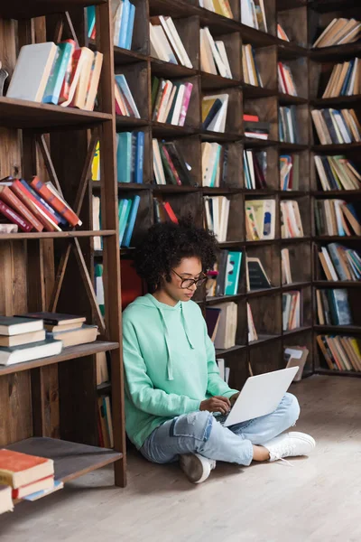 Young african american woman in eyeglasses using laptop while sitting on floor in library — Stock Photo