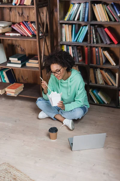 Junge afrikanisch-amerikanische Frau mit Brille sitzt auf dem Boden und hält Essstäbchen in der Nähe von Imbissen in der Bibliothek — Stockfoto