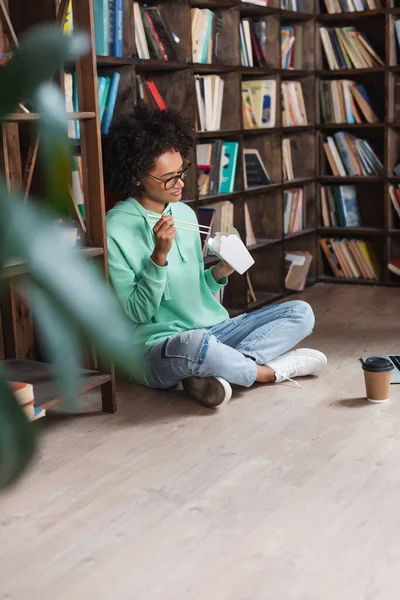 Young african american woman in eyeglasses sitting on floor and holding chopsticks near takeaway food in library — Stock Photo