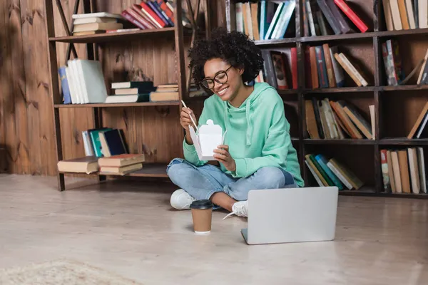 Glückliche afrikanisch-amerikanische Frau mit Brille sitzt auf dem Boden und hält Essstäbchen in der Nähe von Imbissen in der Bibliothek — Stockfoto