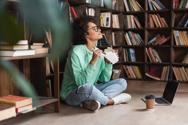 Pleased african american woman in eyeglasses sitting on floor and holding chopsticks near carton box with takeaway food near laptop — Stock Photo