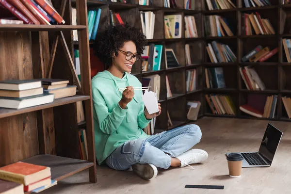 Happy african american woman in eyeglasses sitting on floor and holding chopsticks near carton box with takeaway food near laptop — Stock Photo