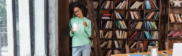 Joven afroamericana mujer en gafas con palillos cerca de caja de cartón con comida para llevar, pancarta - foto de stock