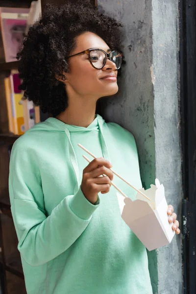 Heureuse femme afro-américaine dans des lunettes tenant des baguettes près de la boîte en carton avec déjeuner à emporter — Photo de stock