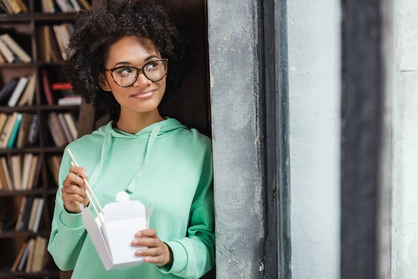 Femme afro-américaine souriante dans des lunettes tenant des baguettes près de la boîte en carton avec déjeuner à emporter — Photo de stock