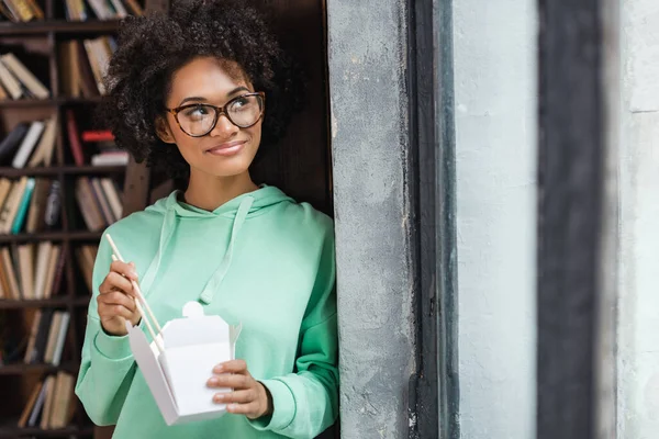 Mujer afroamericana sonriente en gafas con palillos cerca de caja de cartón con almuerzo para llevar - foto de stock