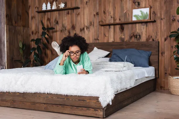 Африканская американка регулирует очки и лежит на кровати, используя смартфон рядом с ноутбуком — стоковое фото