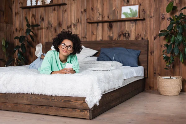 Nachdenkliche Afroamerikanerin mit Brille im Bett liegend, während sie ihr Smartphone in Laptopnähe benutzt — Stockfoto