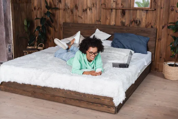 Fröhliche afrikanisch-amerikanische Frau mit Brille, die auf dem Bett liegt, während sie ihr Smartphone in Laptopnähe benutzt — Stockfoto