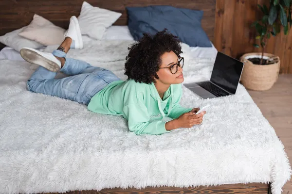 Joven afroamericana mujer en gafas acostado en la cama y la celebración de teléfono inteligente cerca de la computadora portátil - foto de stock