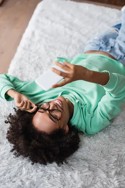 Vue grand angle de femme afro-américaine heureuse dans des lunettes couché sur le lit et de prendre selfie — Photo de stock